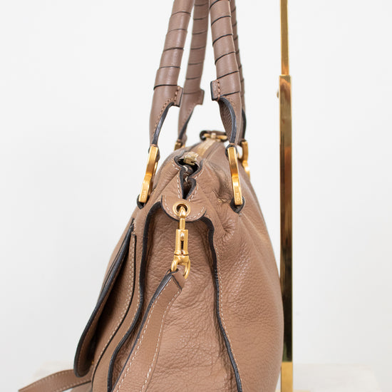 A taupe brown pebbled leather handbag with gold-tone hardware, displayed on a minimalist gold metal stand against a clean white background.