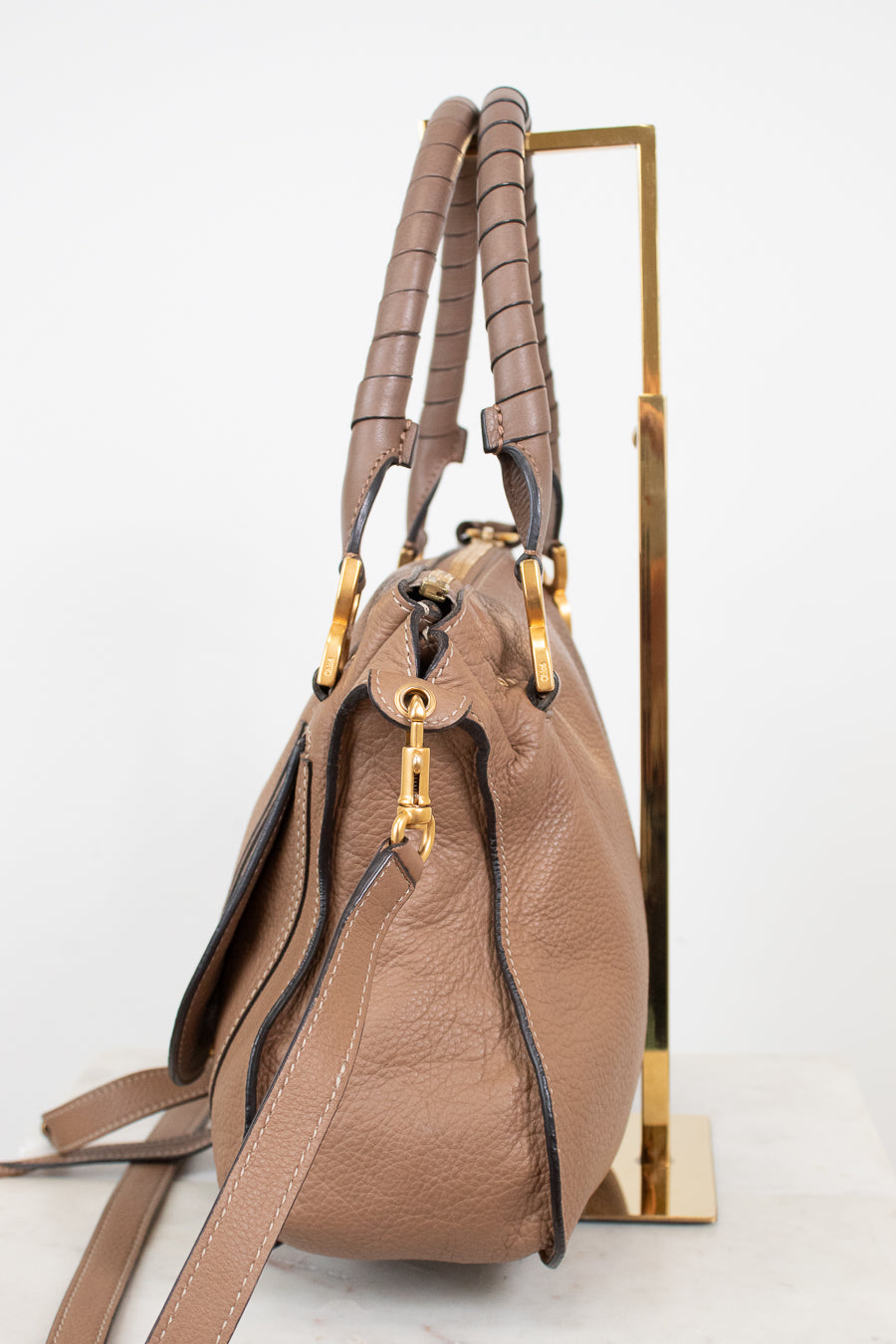 A taupe brown pebbled leather handbag with gold-tone hardware, displayed on a minimalist gold metal stand against a clean white background.