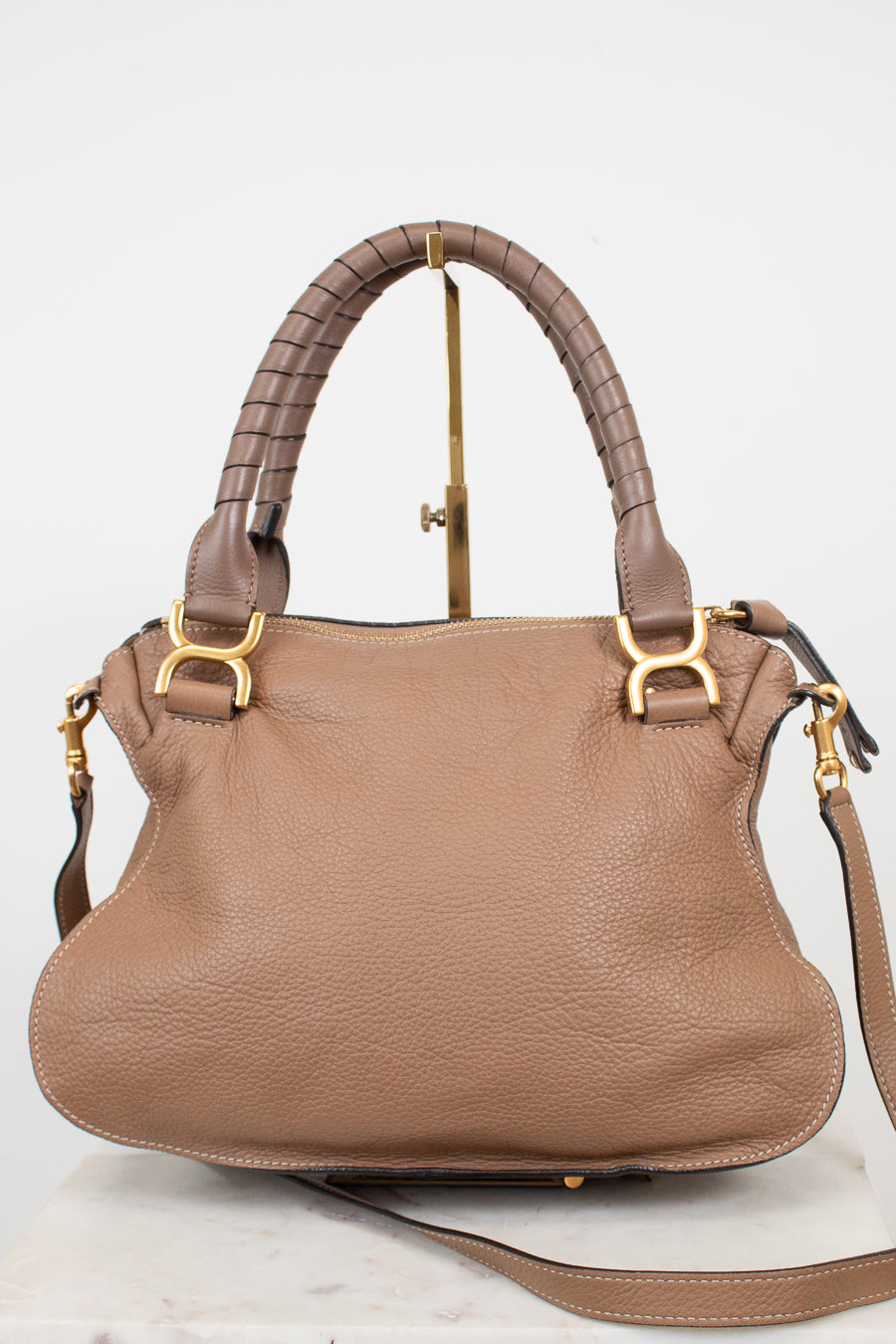 A taupe brown pebbled leather handbag with a braided top handle, gold-tone hardware, and a matching shoulder strap, displayed on a stand against a white background.