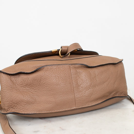 A tan leather shoulder bag with a pebbled texture laying with bottom in view, featuring a curved side edge, a flap closure gold-tone ring, and a thin strap, photographed on a white marble surface against a white background.