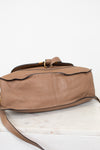 A tan leather shoulder bag with a pebbled texture laying with bottom in view, featuring a curved side edge, a flap closure gold-tone ring, and a thin strap, photographed on a white marble surface against a white background.