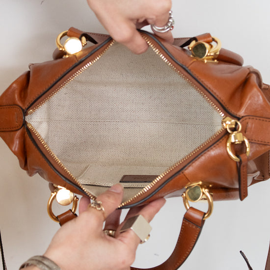 A close-up of a brown leather handbag being opened by two hands, showing the beige mesh interior and gold-tone hardware against a plain white background.
