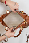 A close-up of a brown leather handbag being opened by two hands, showing the beige mesh interior and gold-tone hardware against a plain white background.
