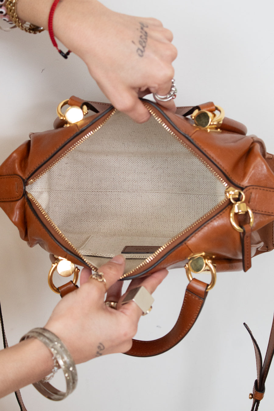 A close-up of a brown leather handbag being opened by two hands, showing the beige mesh interior and gold-tone hardware against a plain white background.