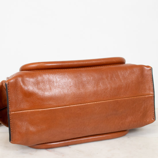 A close-up of a brown leather handbag or satchel showing the side and bottom profile, resting on a light marble surface against a white background.