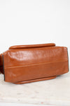 A close-up of a brown leather handbag or satchel showing the side and bottom profile, resting on a light marble surface against a white background.