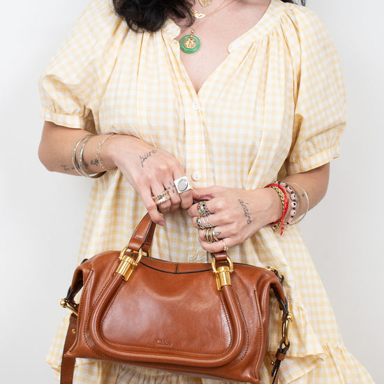 A woman wearing a yellow and white gingham dress holds a brown leather handbag with gold hardware against a plain light background.