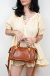 A woman wearing a yellow and white gingham dress holds a brown leather handbag with gold hardware against a plain light background.