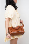 A woman wearing a yellow and white gingham dress carries a brown leather handbag with a top handle and shoulder strap against a plain light gray background.