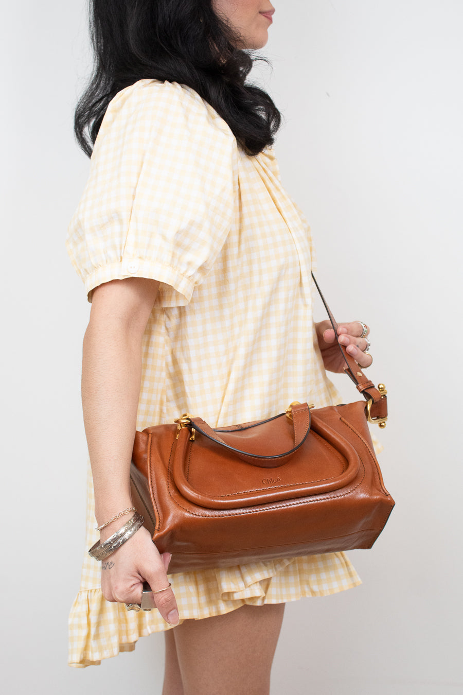 A woman wearing a yellow and white gingham dress carries a brown leather handbag with a top handle and shoulder strap against a plain light gray background.