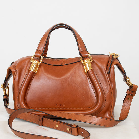 A brown leather handbag with gold-tone hardware, featuring dual top handles and a detachable shoulder strap, displayed on a white surface against a white background.