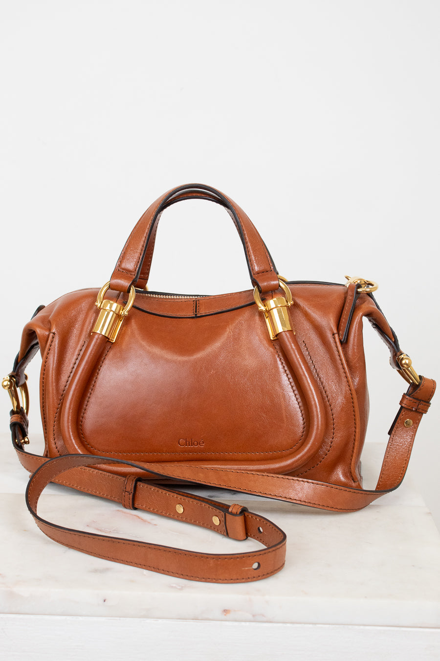 A brown leather handbag with gold-tone hardware, featuring dual top handles and a detachable shoulder strap, displayed on a white surface against a white background.