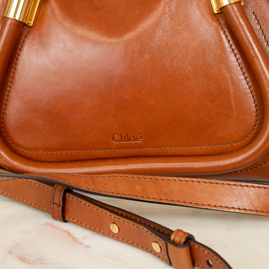 A close-up of a brown leather handbag with gold-tone hardware and a visible 'Chloé' logo on the front, resting on a light marble surface with part of the matching strap visible.