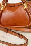 A close-up of a brown leather handbag with gold-tone hardware and a visible 'Chloé' logo on the front, resting on a light marble surface with part of the matching strap visible.