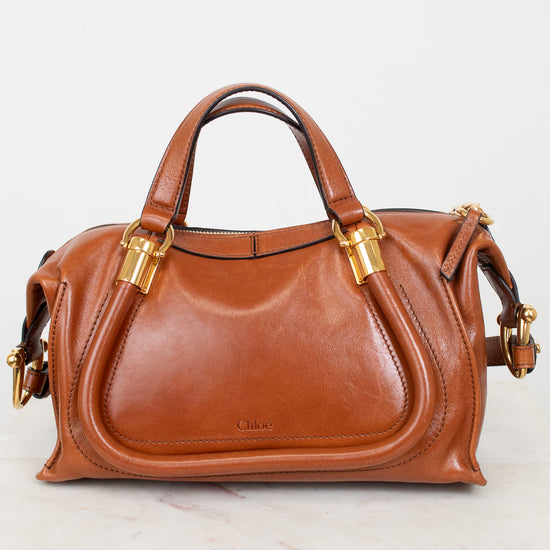 A tan leather handbag with gold-tone hardware, featuring two top handles and a curved front flap, displayed on a light marble surface against a white background.