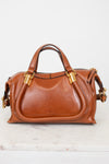 A tan leather handbag with gold-tone hardware, featuring two top handles and a curved front flap, displayed on a light marble surface against a white background.