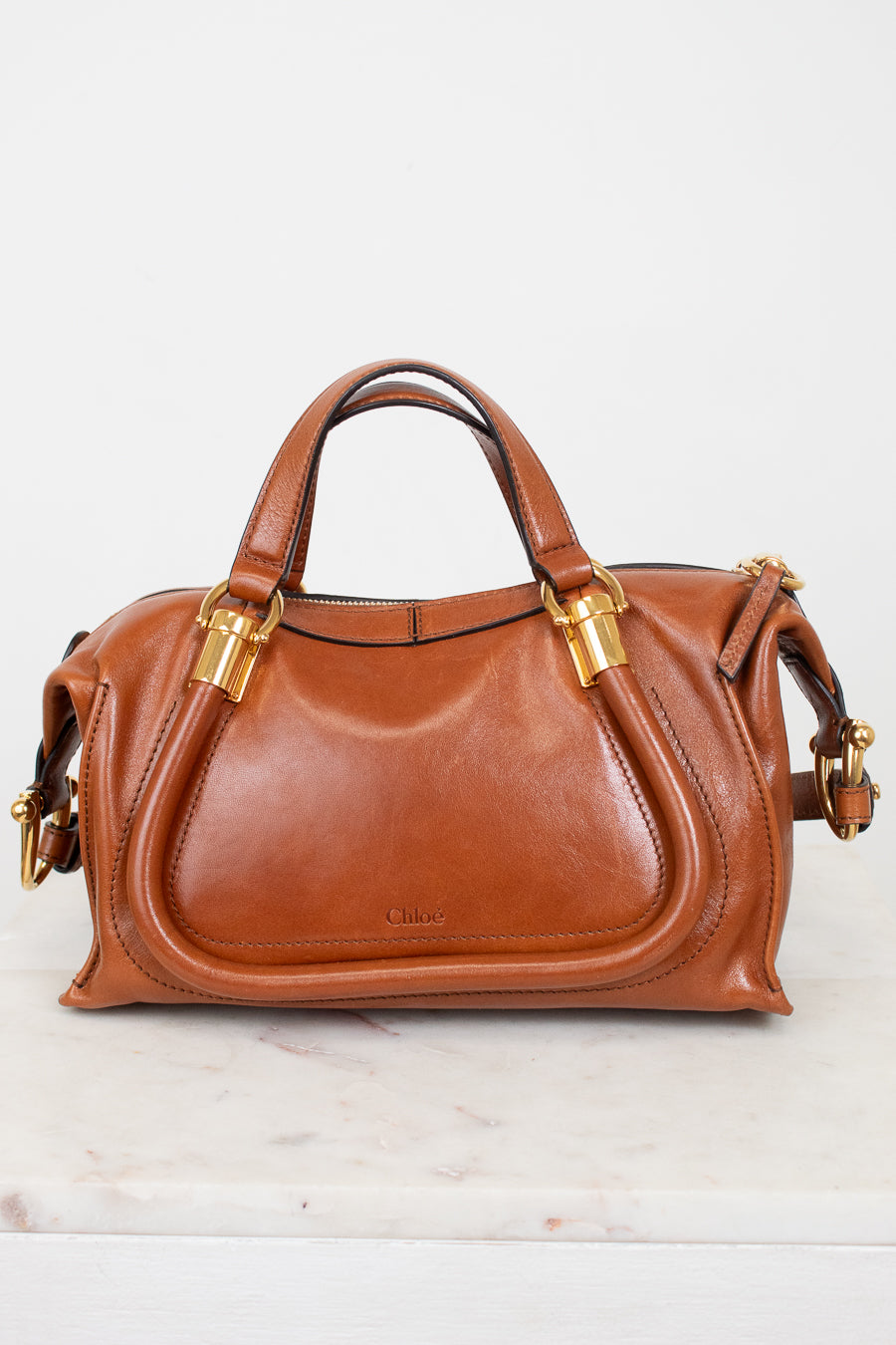 A tan leather handbag with gold-tone hardware, featuring two top handles and a curved front flap, displayed on a light marble surface against a white background.