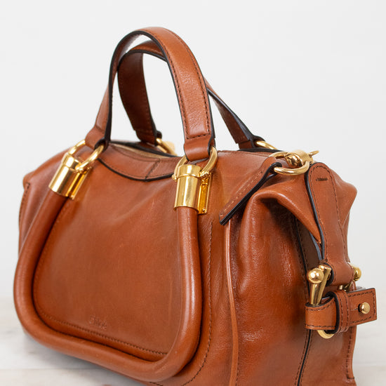 A structured cognac brown leather handbag with gold-tone hardware, dual top handles, and a side strap detail, photographed on a light marble surface against a white background.