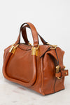 A structured cognac brown leather handbag with gold-tone hardware, dual top handles, and a side strap detail, photographed on a light marble surface against a white background.