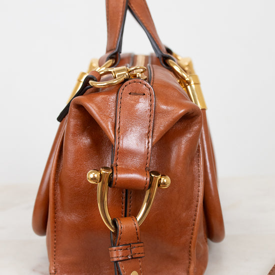 A brown leather handbag with gold-tone hardware, featuring a structured top handle and a long shoulder strap, photographed on a light marble surface against a white background.