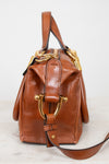 A brown leather handbag with gold-tone hardware, featuring a structured top handle and a long shoulder strap, photographed on a light marble surface against a white background.