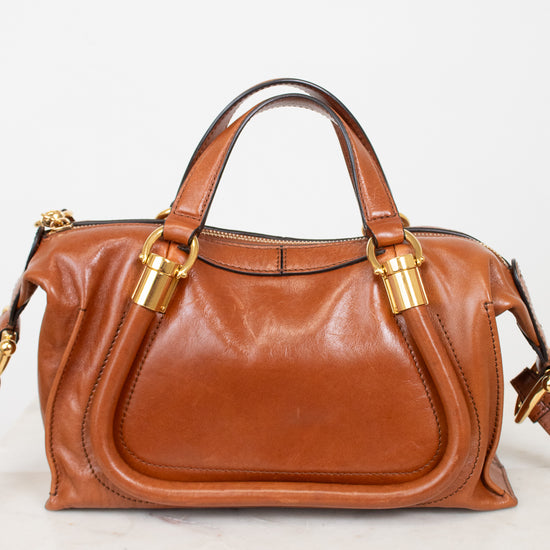 A brown leather handbag with gold-tone hardware, featuring dual top handles and a front slip pocket, displayed on a light marble surface against a white background.