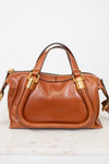 A brown leather handbag with gold-tone hardware, featuring dual top handles and a front slip pocket, displayed on a light marble surface against a white background.