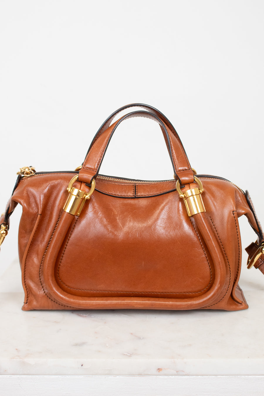 A brown leather handbag with gold-tone hardware, featuring dual top handles and a front slip pocket, displayed on a light marble surface against a white background.