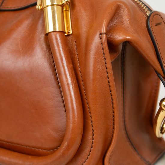 A close-up of a brown leather handbag with gold-tone hardware, showing the side profile, stitching, and a curved leather detail.