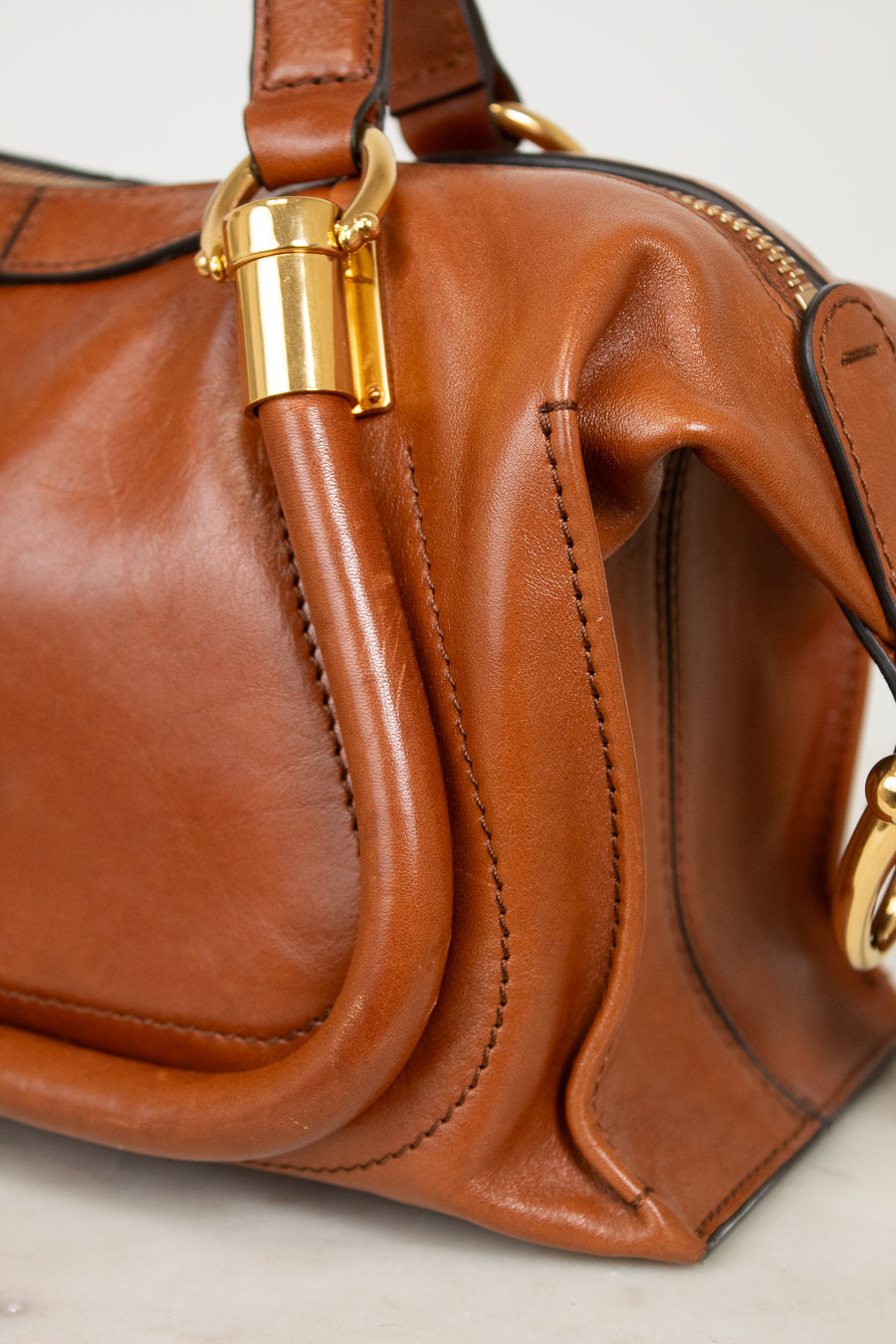 A close-up of a brown leather handbag with gold-tone hardware, showing the side profile, stitching, and a curved leather detail.