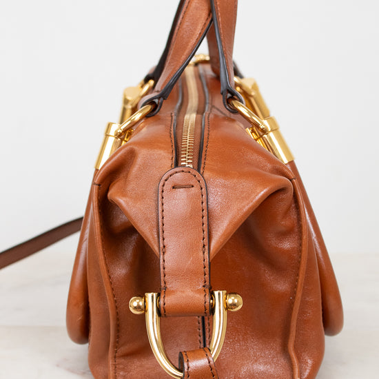 A brown leather handbag with gold-tone hardware, featuring a structured shape, a top handle, a front flap with a buckle strap, and a zip-top closure, photographed against a light background.