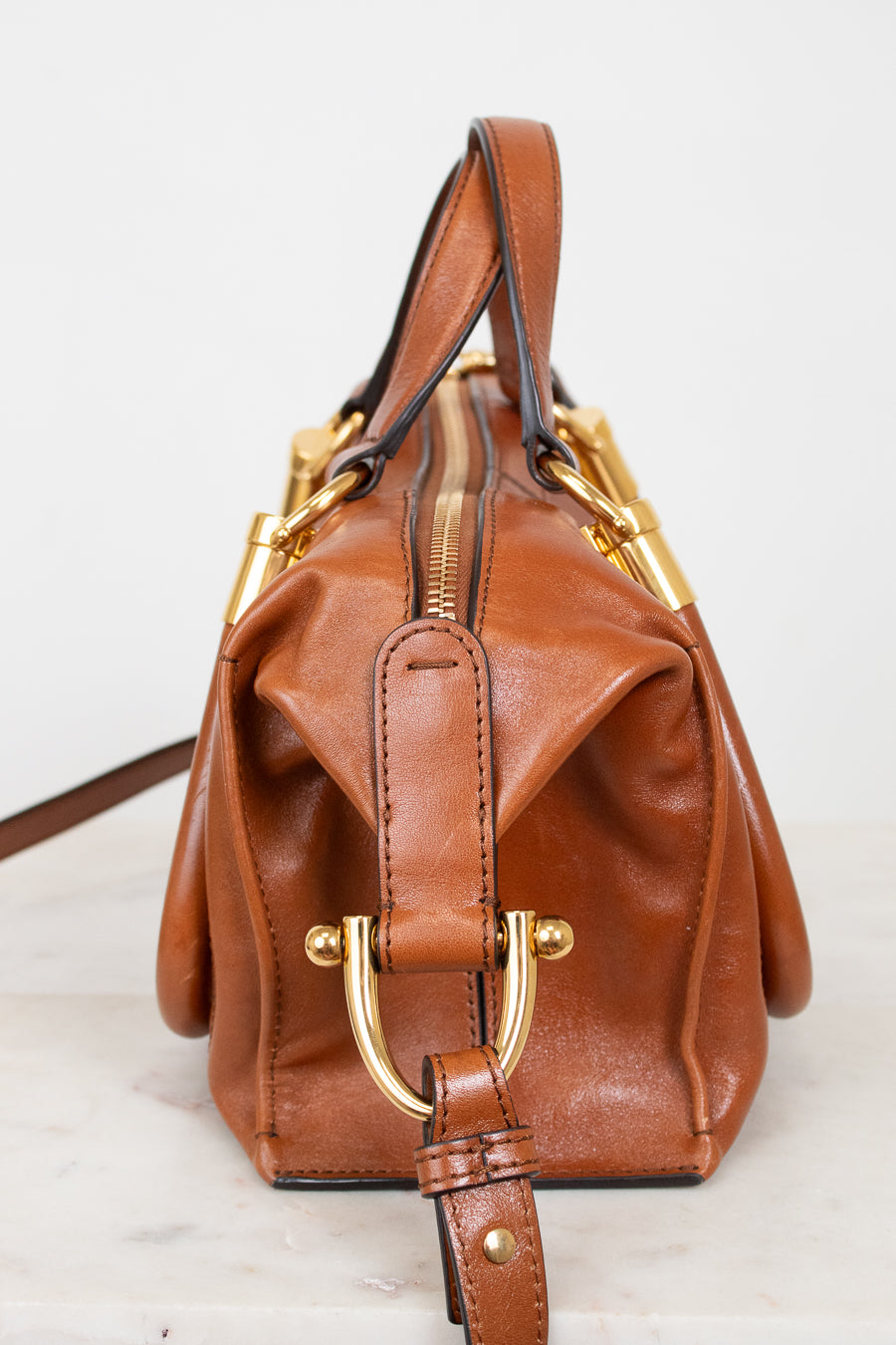 A brown leather handbag with gold-tone hardware, featuring a structured shape, a top handle, a front flap with a buckle strap, and a zip-top closure, photographed against a light background.