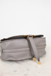 A light gray pebbled leather shoulder bag with gold-tone hardware, resting on a white marble surface against a white background.