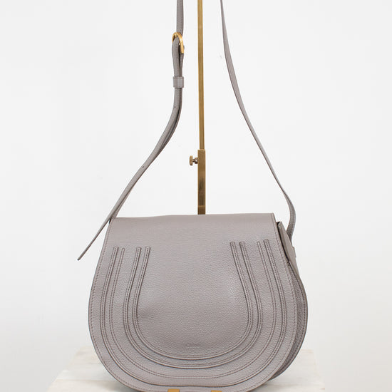 A light gray leather shoulder bag with a curved flap, vertical stitched details, and gold-tone hardware, displayed on a white marble block against a white background.