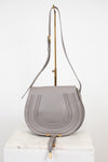 A light gray leather shoulder bag with a curved flap, vertical stitched details, and gold-tone hardware, displayed on a white marble block against a white background.