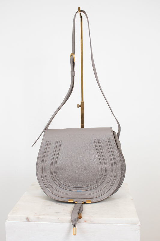 A light gray leather shoulder bag with a curved flap, vertical stitched details, and gold-tone hardware, displayed on a white marble block against a white background.