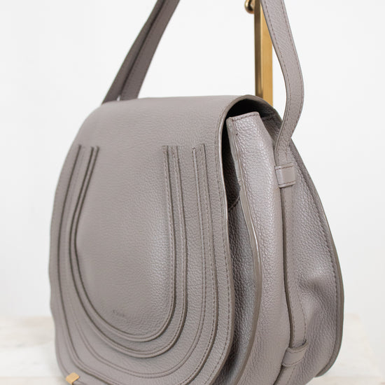 A light gray pebbled leather shoulder bag with a front flap, vertical stitched detailing, and a gold-tone metal stand visible behind it, set against a clean white background.