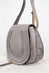 A light gray pebbled leather shoulder bag with a front flap, vertical stitched detailing, and a gold-tone metal stand visible behind it, set against a clean white background.