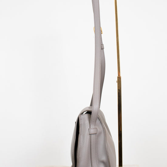 A light gray leather shoulder bag with gold-tone hardware, displayed on a white marble surface next to a gold metal stand against a white background.