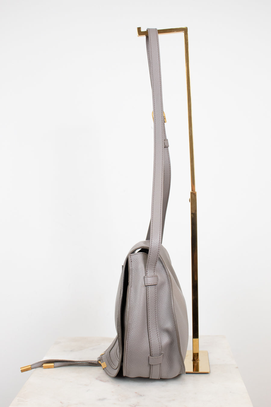 A light gray leather shoulder bag with gold-tone hardware, displayed on a white marble surface next to a gold metal stand against a white background.