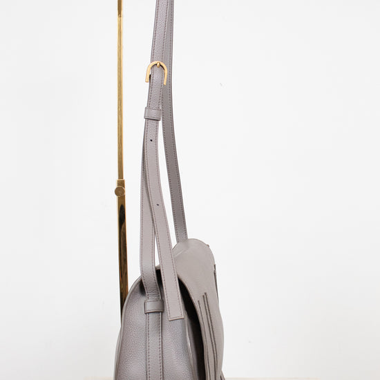 A light gray leather shoulder bag with gold-tone hardware, shown hanging from a gold display stand against a white studio background.