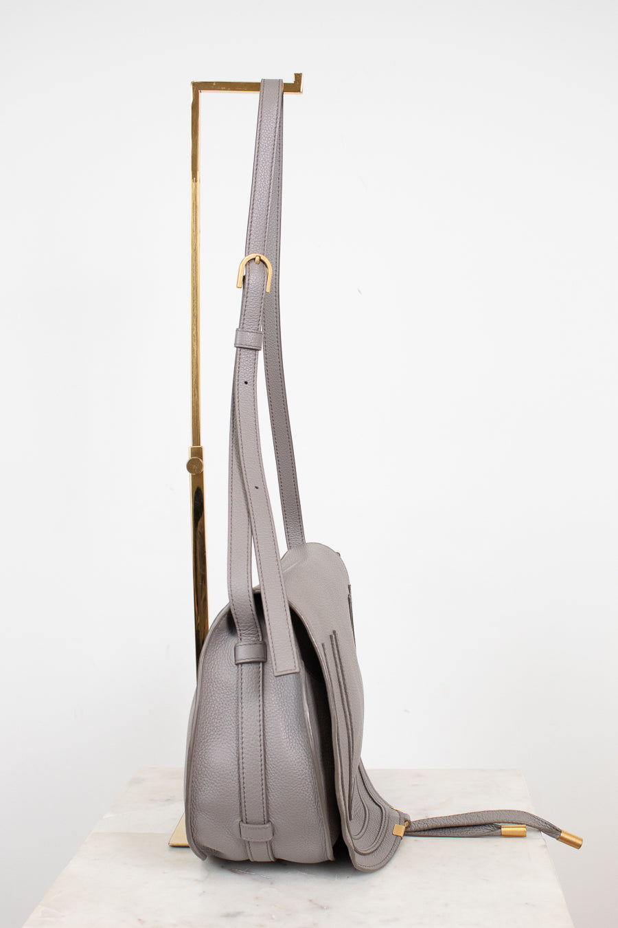 A light gray leather shoulder bag with gold-tone hardware, shown hanging from a gold display stand against a white studio background.