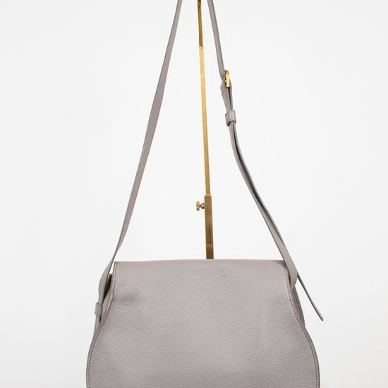 A light gray leather shoulder bag with a curved bottom and long shoulder strap, displayed on a gold stand against a white background.