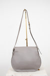 A light gray leather shoulder bag with a curved bottom and long shoulder strap, displayed on a gold stand against a white background.