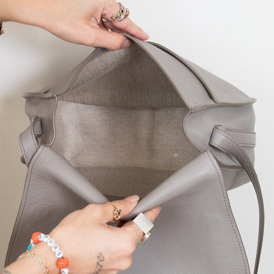 A close-up of a person's hands opening a large, structured gray pebbled leather bag, showing the interior gray lining and strap attachment.