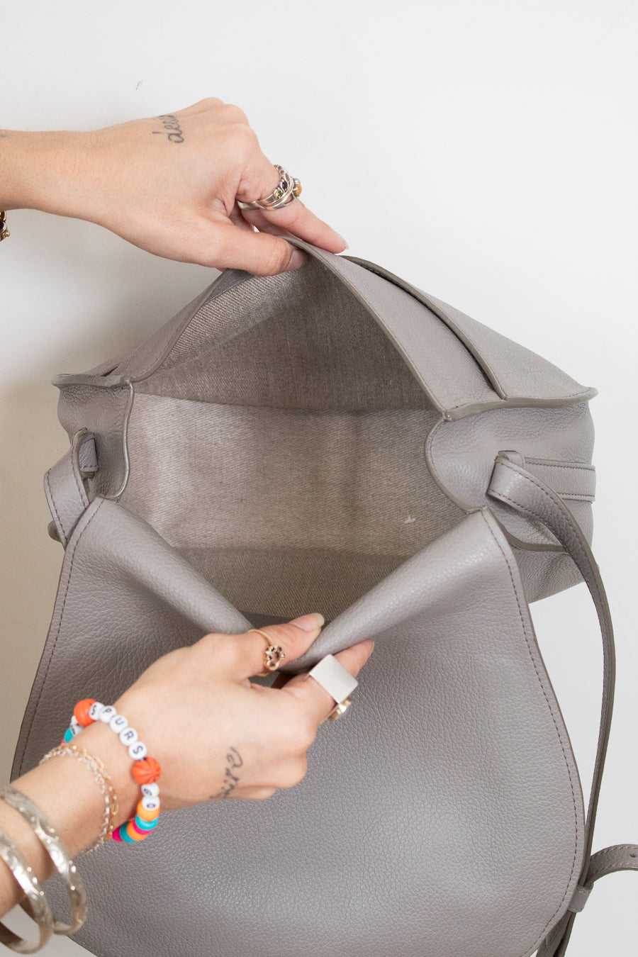 A close-up of a person's hands opening a large, structured gray pebbled leather bag, showing the interior gray lining and strap attachment.