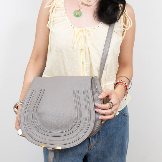 A woman wearing a cream ruffled top and blue jeans carries a light gray leather shoulder bag with gold-tone hardware against a plain light gray background.