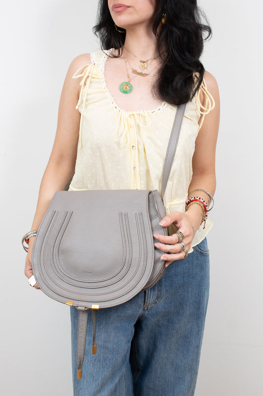 A woman wearing a cream ruffled top and blue jeans carries a light gray leather shoulder bag with gold-tone hardware against a plain light gray background.