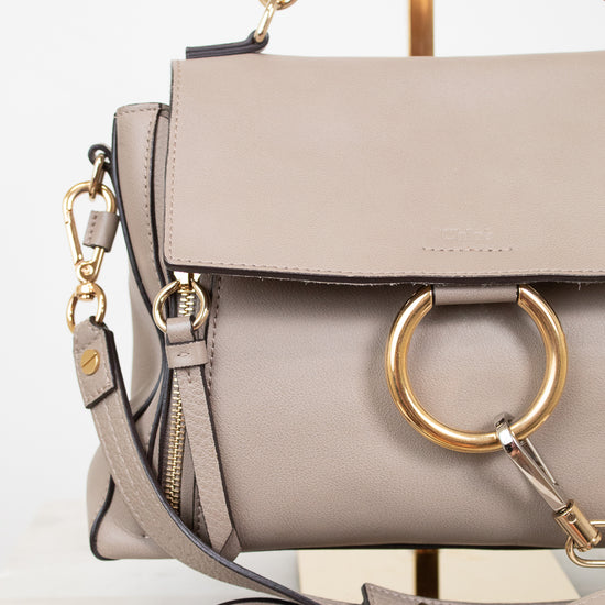 A taupe leather handbag with a large gold-tone O-ring on the front flap, a matching shoulder strap, and gold-tone hardware, displayed on a stand against a white background.