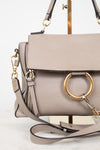 A taupe leather handbag with a large gold-tone O-ring on the front flap, a matching shoulder strap, and gold-tone hardware, displayed on a stand against a white background.